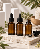 Three amber glass dropper bottles with black caps are arranged on a light stone slab, with one bottle open and a glass dropper lying beside it. White rectangular boxes, woven decor, and green leaves are softly blurred in the background.