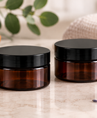 Two amber PET cosmetic jars with black screw-on lids sit on a light marble surface, styled with eucalyptus leaves, a bowl of white scrub, a folded beige towel, and a small bowl of lavender in the background.