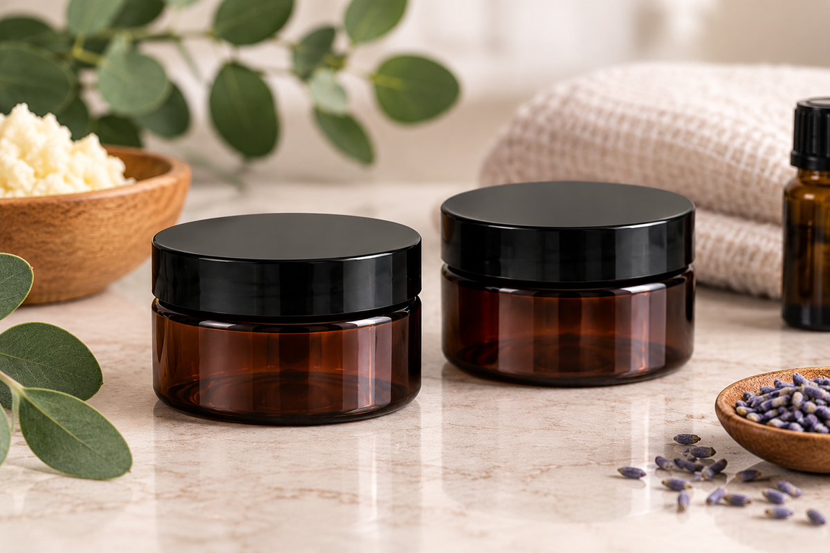 Two amber PET cosmetic jars with black screw-on lids sit on a light marble surface, styled with eucalyptus leaves, a bowl of white scrub, a folded beige towel, and a small bowl of lavender in the background.