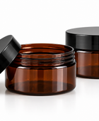 Two empty amber PET cosmetic jars with black screw-on lids on a clean white background, with one jar open and its lid leaning against it.