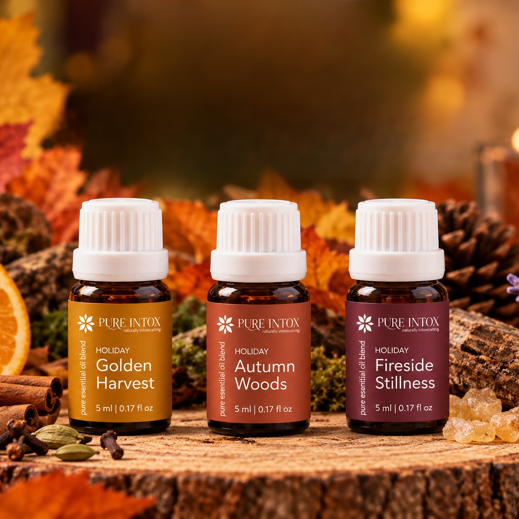 Three Pure Intox essential oil bottles from the Holiday Limited Autumn Collection are displayed on a rustic wood slice surrounded by autumn leaves, orange slices, cinnamon, and a candle. The bottles are labeled Golden Harvest, Autumn Woods, and Fireside Stillness.