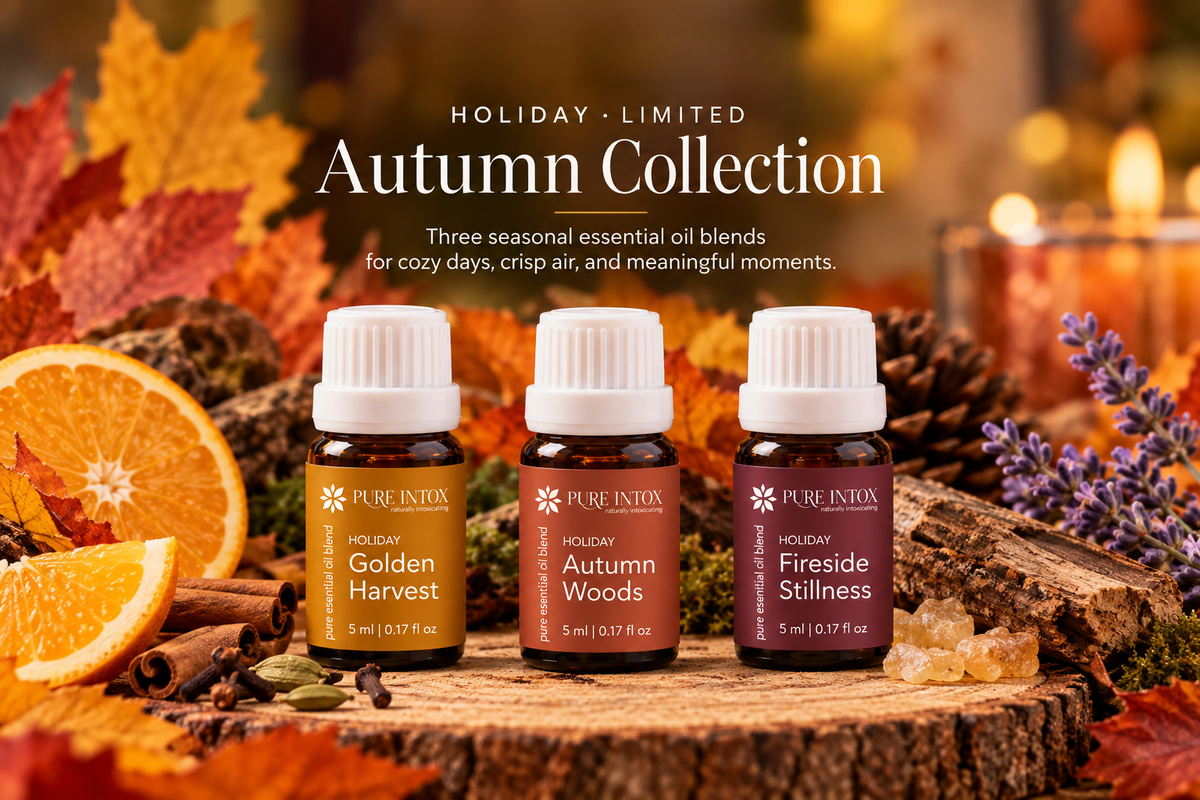 Three Pure Intox essential oil bottles from the Holiday Limited Autumn Collection are displayed on a rustic wood slice surrounded by autumn leaves, orange slices, cinnamon, and a candle. The bottles are labeled Golden Harvest, Autumn Woods, and Fireside Stillness.