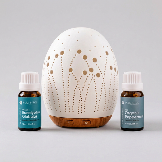 A white ceramic diffuser with laser-cut geometric patterns and a wooden base, flanked by two Pure Inton essential oil bottles labeled Organic Eucalyptus Globulus and Organic Peppermint on a white background.