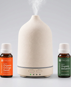 A Pure Intox essential oil diffuser set featuring a textured cream-colored diffuser in the center, emitting a plume of steam, flanked by two small amber bottles of Pure Inton essential oils: Organic Orange Sweet on the left with an orange label, and Organic Balsam Fir on the right with a green label.