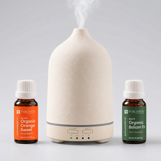 A Pure Intox essential oil diffuser set featuring a textured cream-colored diffuser in the center, emitting a plume of steam, flanked by two small amber bottles of Pure Inton essential oils: Organic Orange Sweet on the left with an orange label, and Organic Balsam Fir on the right with a green label.