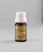 A bottle of Pure Intox 'Herb Coriander Seed' essential oil placed on a rustic wooden board surrounded by fresh coriander leaves, loose seeds, and a wooden bowl, with a blurred traditional mortar and pestle in the background.