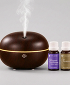 A dark wood-look essential oil diffuser with a glowing light band and misting at the top, shown next to two Pure Intoxessential oil bottles labeled Organic Lavender and Frankincense on a white background.