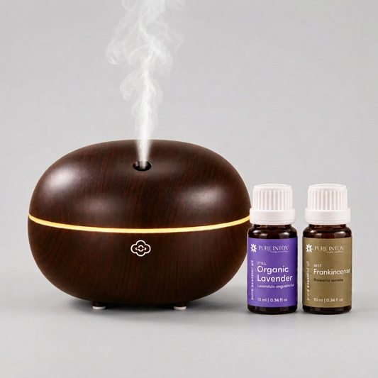 A dark wood-look essential oil diffuser with a glowing light band and misting at the top, shown next to two Pure Intoxessential oil bottles labeled Organic Lavender and Frankincense on a white background.