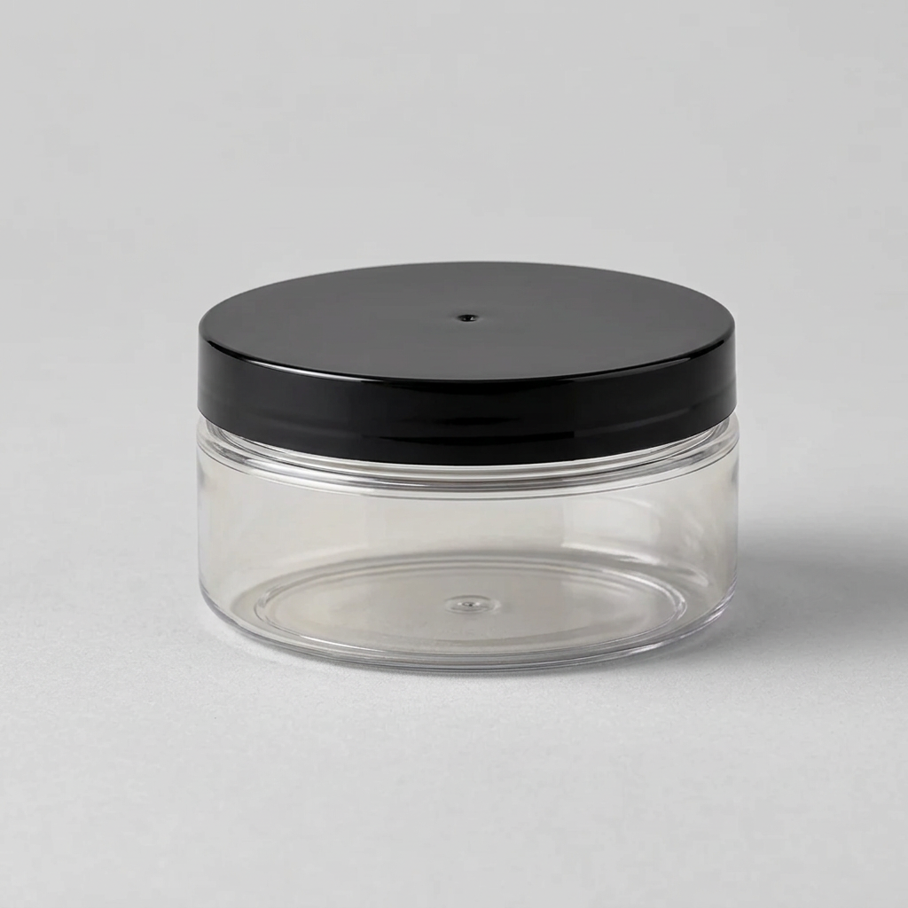 A clear round cosmetic jar with a glossy black lid, set on a marble surface with folded white towels, green leaves, and a soap dish in the background.