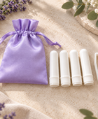 A lavender satin drawstring pouch, four white lip balm tubes, and a pair of white tweezers arranged on a beige textured surface with decorative lavender and greenery.
