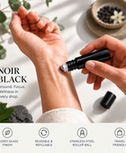A close-up of a person's hands applying cream from a black roll-on bottle to the back of their hand. The text reads 'NOIR BLACK' with supporting copy about the glossy glass finish, reusable refillable design, stainless steel roller ball, and travel-friendly packaging.
