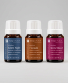 Three small amber glass essential oil bottles with white ribbed caps on a light gray background. The labels read PURE INTOX and show three holiday scent options: Holiday Silent Night in blue, Holiday Fireside in brown, and Holiday Winter Bloom in magenta.