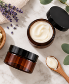 Two amber cosmetic jars with black lids on a light marble surface, one open to show a white cream and one closed. A wooden spoon holds more cream, with lavender sprigs, eucalyptus leaves, and a small bowl of pale yellow solid product arranged around them.