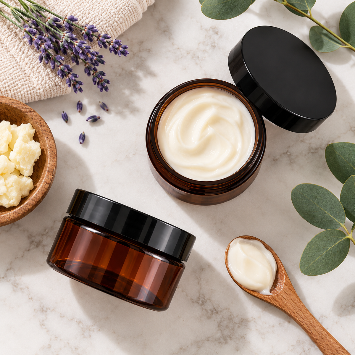 Two amber cosmetic jars with black lids on a light marble surface, one open to show a white cream and one closed. A wooden spoon holds more cream, with lavender sprigs, eucalyptus leaves, and a small bowl of pale yellow solid product arranged around them.