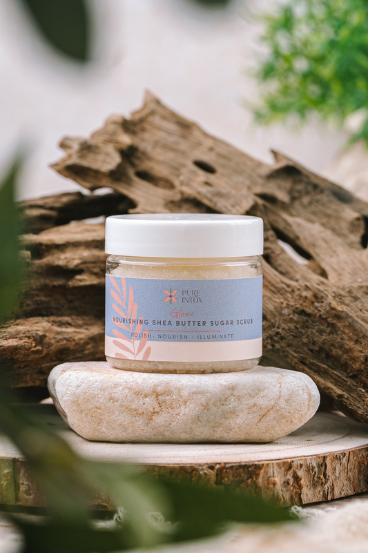 A jar of Pure Intox Organic Guatemalan Shea Butter Sugar Scrub sits on a smooth white stone, with a rustic piece of driftwood and blurred green foliage in the background.