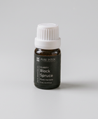 A small black glass bottle of Pure Intox Clarity Black Spruce essential oil with a white ribbed cap, styled on a stone with driftwood, pine needles, and burlap in an outdoor rustic setting.