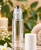 A clear glass roller bottle with a silver ball top and a separate white cap, styled on a wooden surface with green leaves, white flowers, a lit candle, and a soft beige textile in the background.