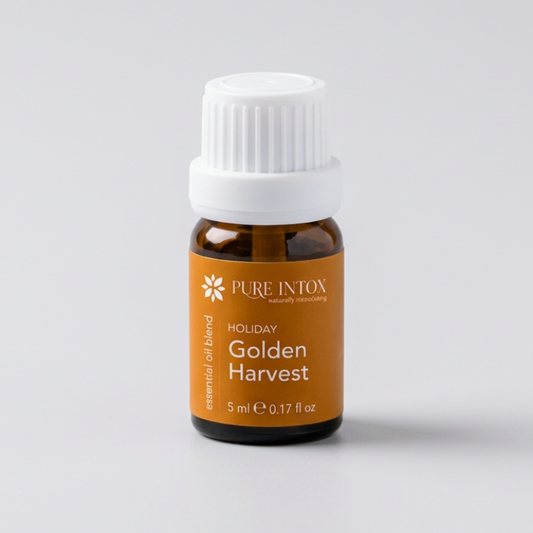A bottle of Pure Intox  Golden Harvest essential oil blend with a white cap and amber label, styled on a stone surrounded by autumn leaves, wheat, and pumpkins against a rustic wood background.