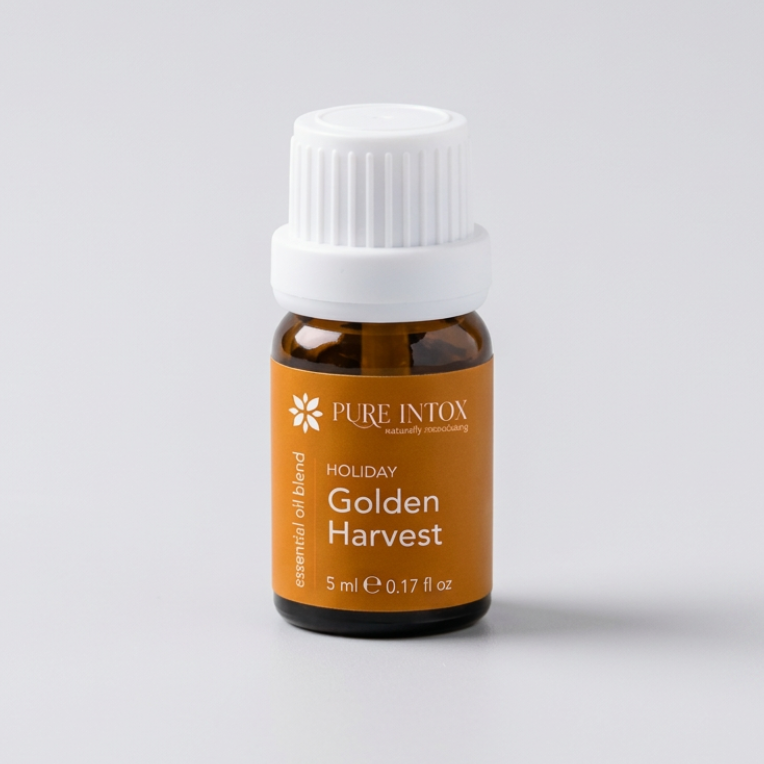A bottle of Pure Intox  Golden Harvest essential oil blend with a white cap and amber label, styled on a stone surrounded by autumn leaves, wheat, and pumpkins against a rustic wood background.